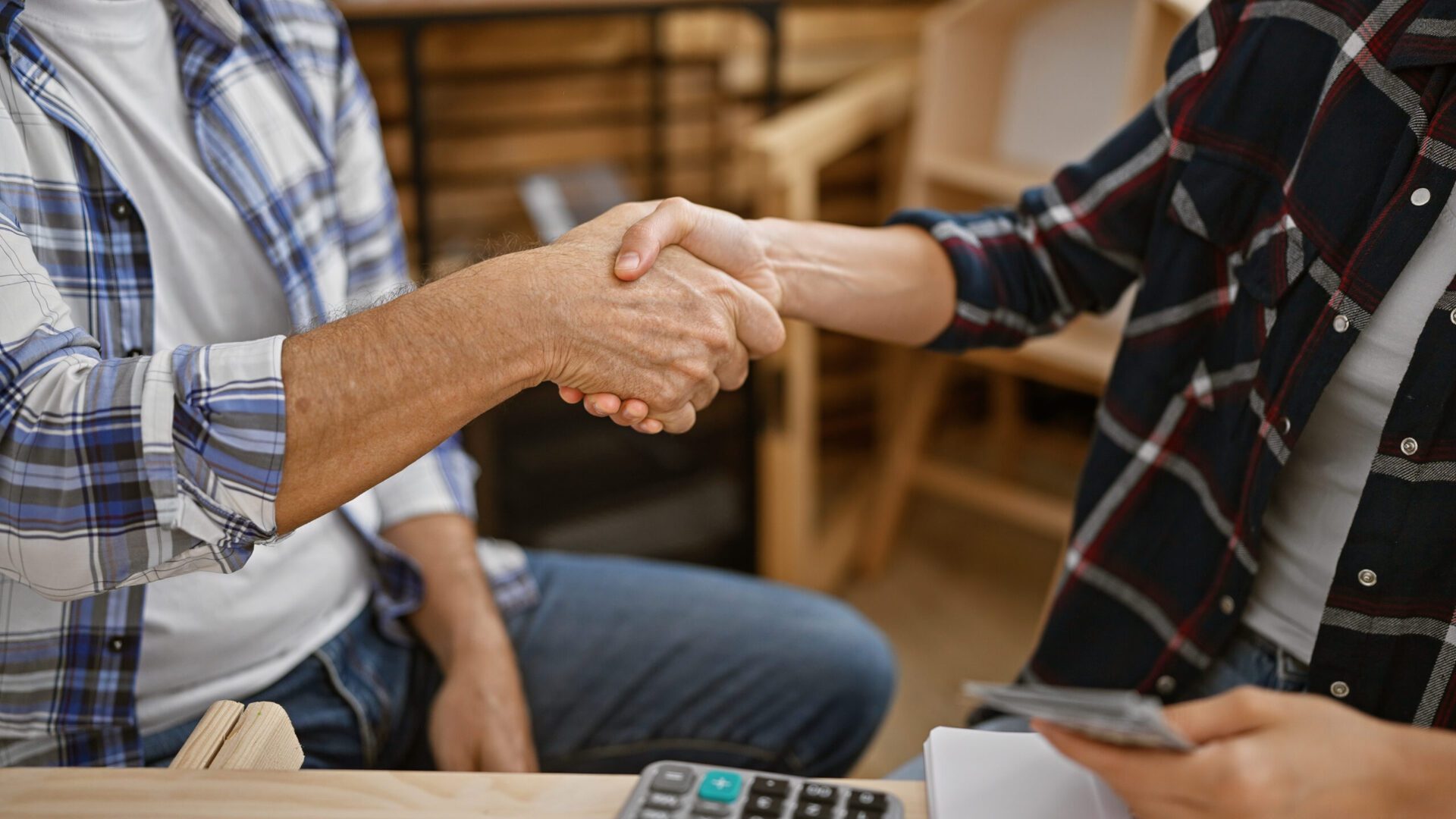 two hearty carpenters, handing over dollars, seal a woodwork agreement with a firm handshake at their cozy indoor carpentry workplace